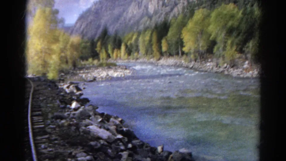 From a moving train, the camera looks along tracks beside a river bordered by rocks, trees, and distant mountains.