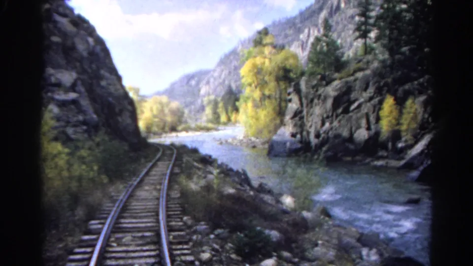 Railroad tracks curve along a river through a rocky canyon, with trees and mountain slopes visible ahead.