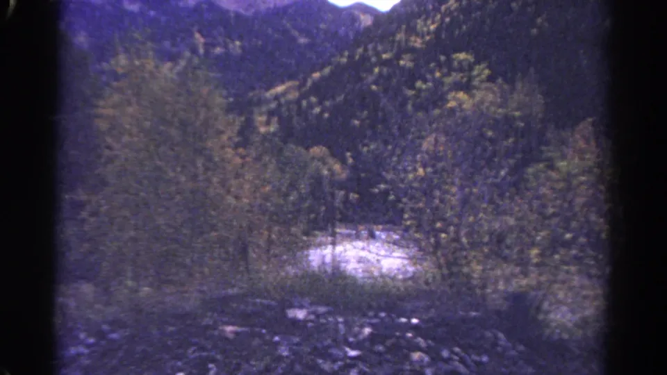 Faded 8mm frame showing a pale rushing stream cutting through a wooded mountain valley with autumn foliage and evergreen slopes.