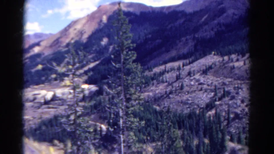1967 8mm home-movie view of evergreen trees, rugged slopes, and a distant mountain valley under bright sky.