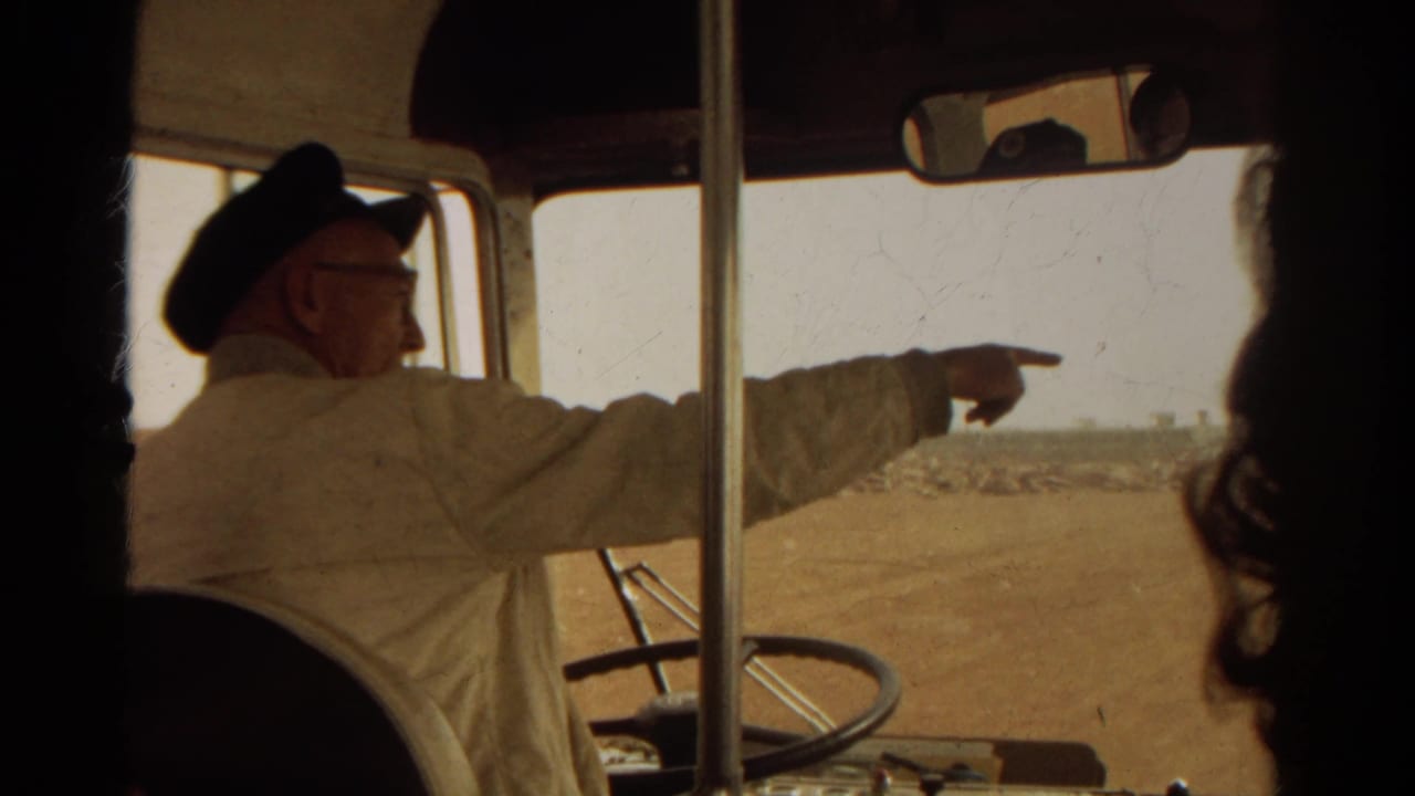 Uniformed man in a dark cap points out a bus window toward a dry landscape.
