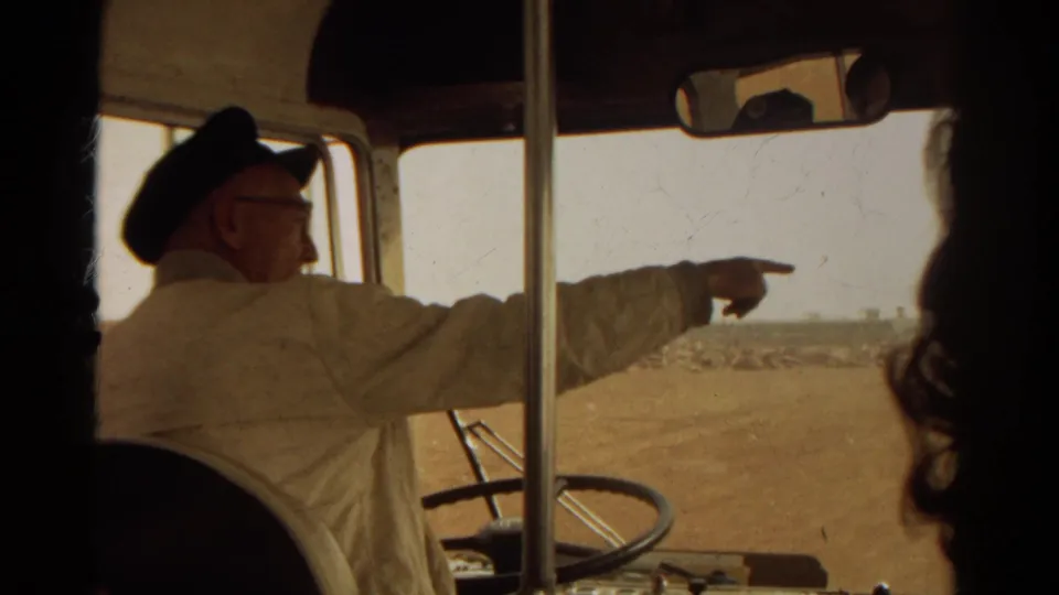1967 8mm frame showing a uniformed man with glasses pointing from the front of a bus toward an arid roadside view.