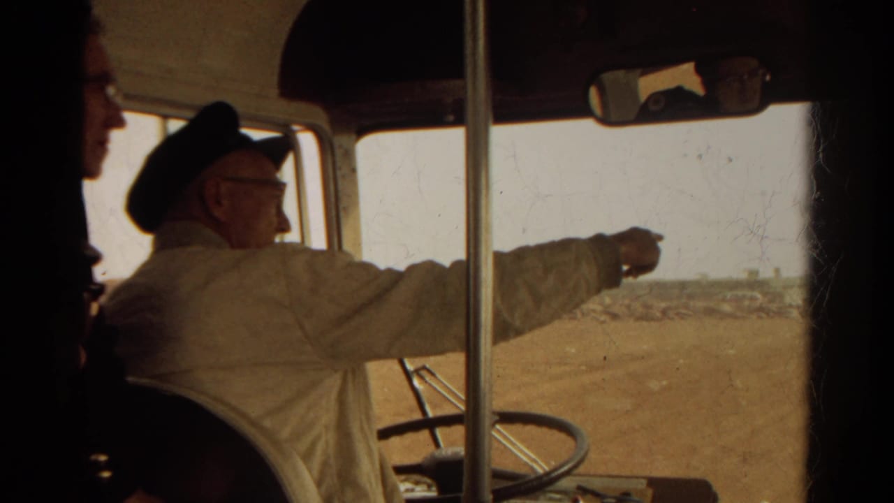 Man in a light uniform points through a dusty bus window as a passenger silhouette crosses the frame.