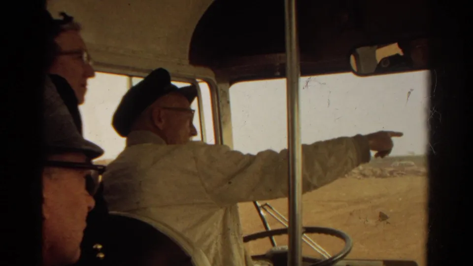 Inside a 1967 bus, seated passengers appear in the foreground while a uniformed man points toward the dry land outside.
