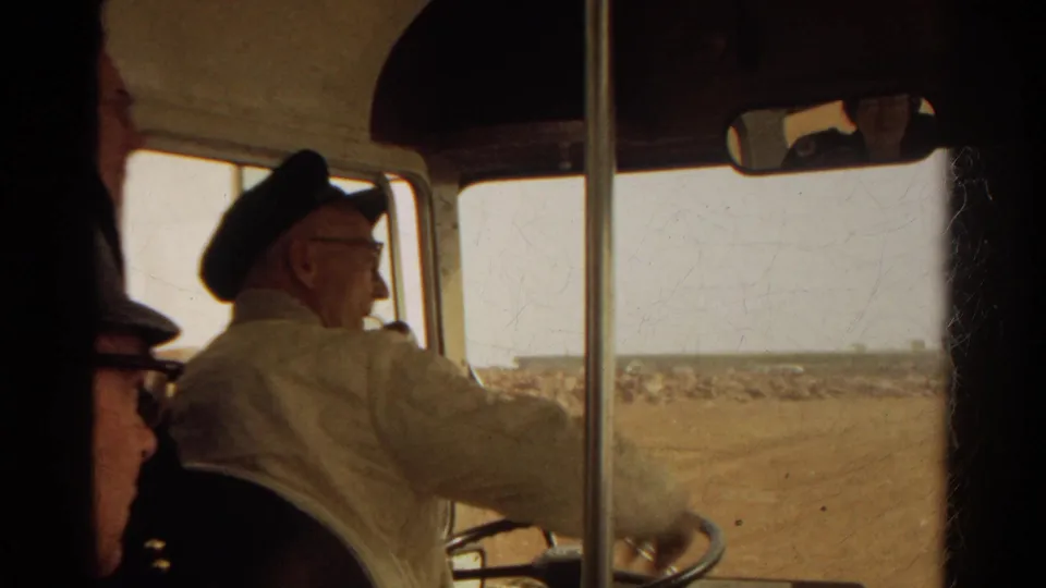 The man in a light uniform sits at the front of the bus near the steering wheel as a dry roadside landscape passes outside.