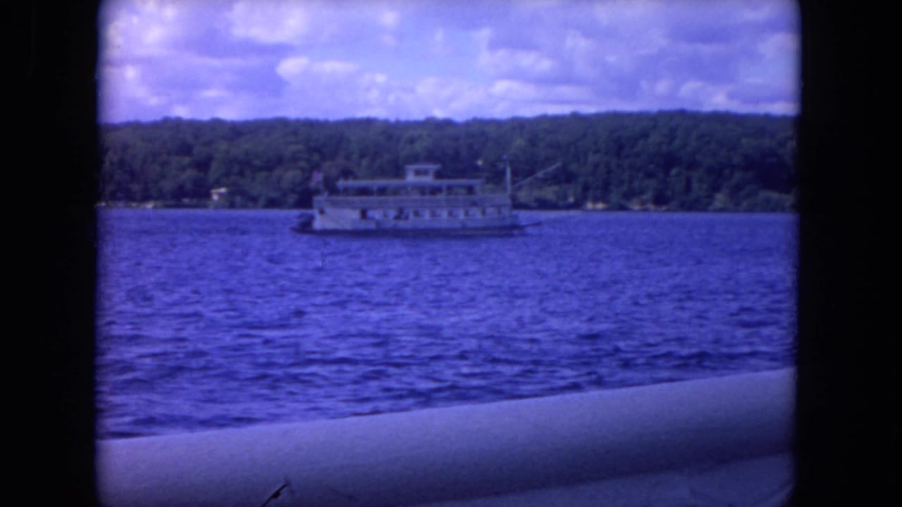 Distant white excursion boat on blue lake water with wooded shoreline and cloudy sky.