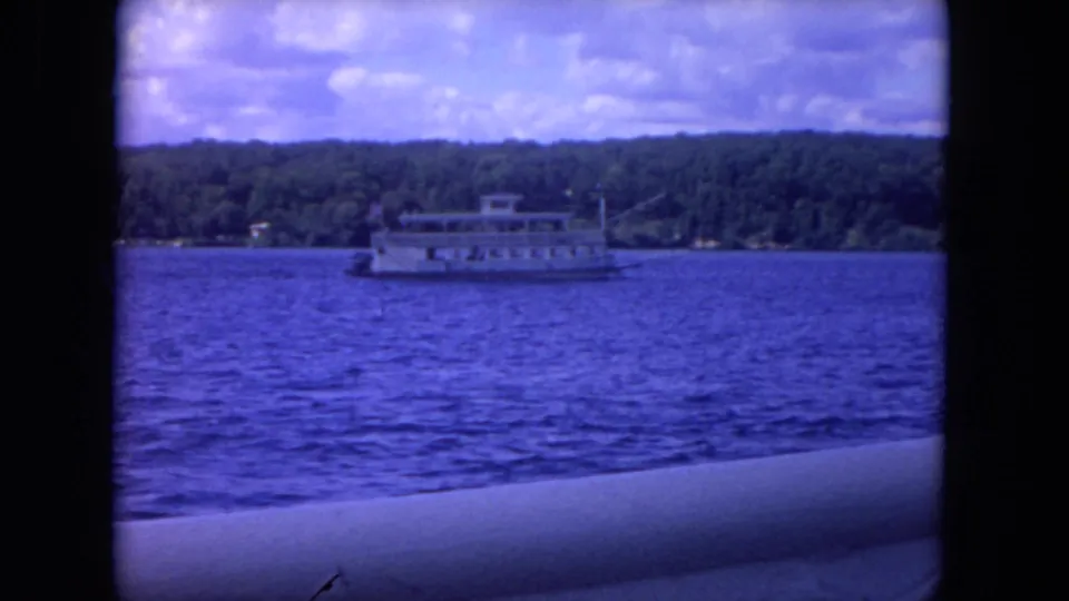 1972 8mm home-movie frame showing a distant white paddlewheel-style boat crossing a lake near Chicago, with trees and clouds behind it.