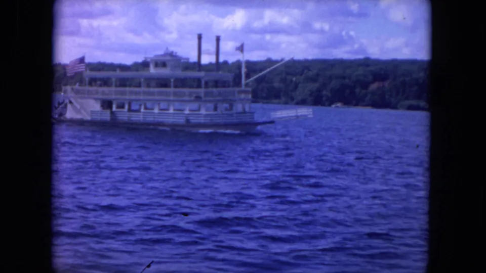 Closer 1972 8mm view of a white paddlewheel-style boat moving across lake water, with flags, smokestacks, and wooded shore behind it.