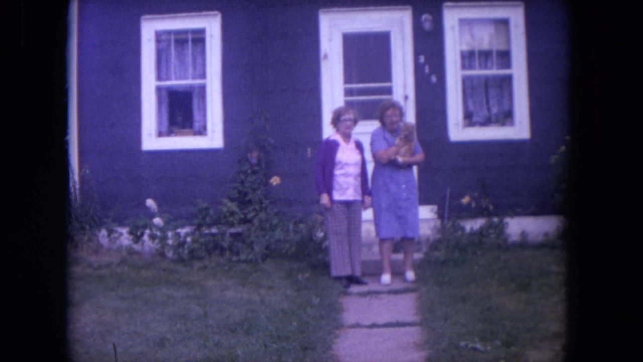 Two women stand on a front path outside a dark blue house while one holds a small dog.