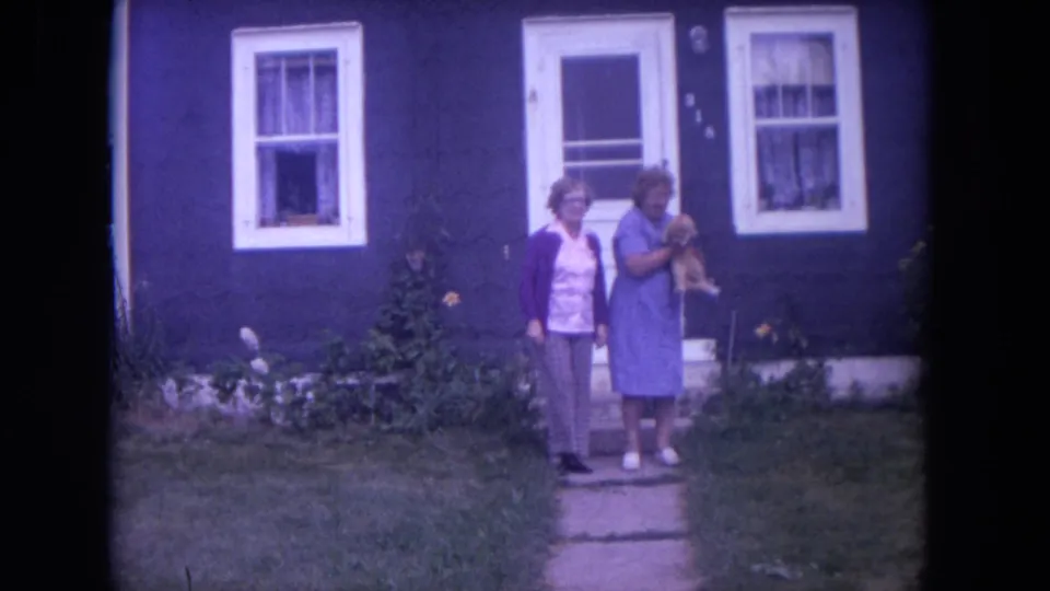 1972 Chicago home-movie frame showing two women on a front path, one in a blue dress holding a small dog near white-trimmed windows.
