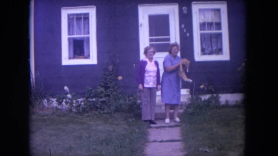 1972 Chicago 8mm frame of two women on a stone path, with the woman in a blue dress lifting a small dog outside a blue house.