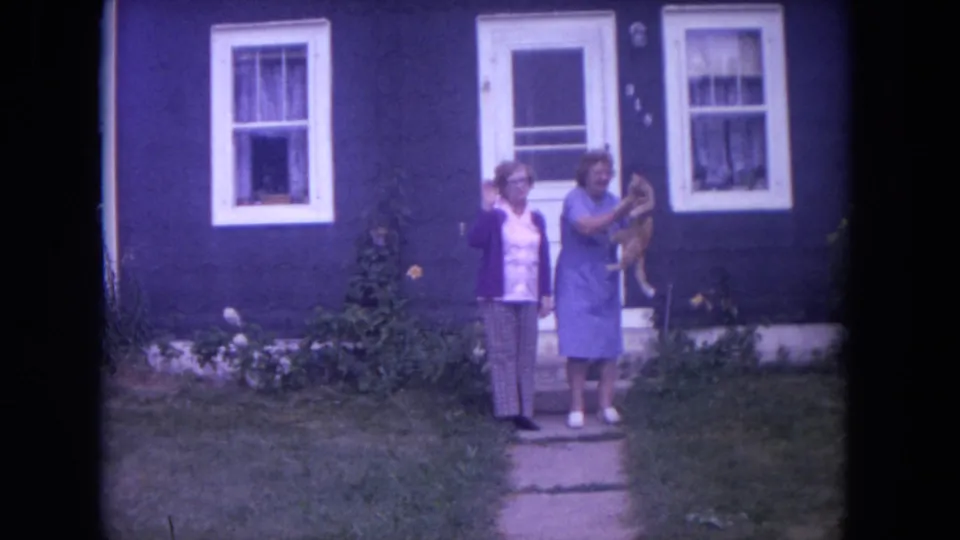 Final 1972 Chicago home-movie frame with two women outside a blue house, one raising a small dog beside the doorway and windows.