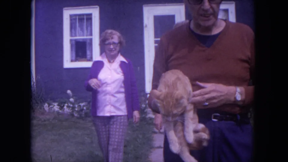 A woman in a purple cardigan and patterned pants walks forward while the man and orange cat fill the right side of the 1972 home-movie frame.