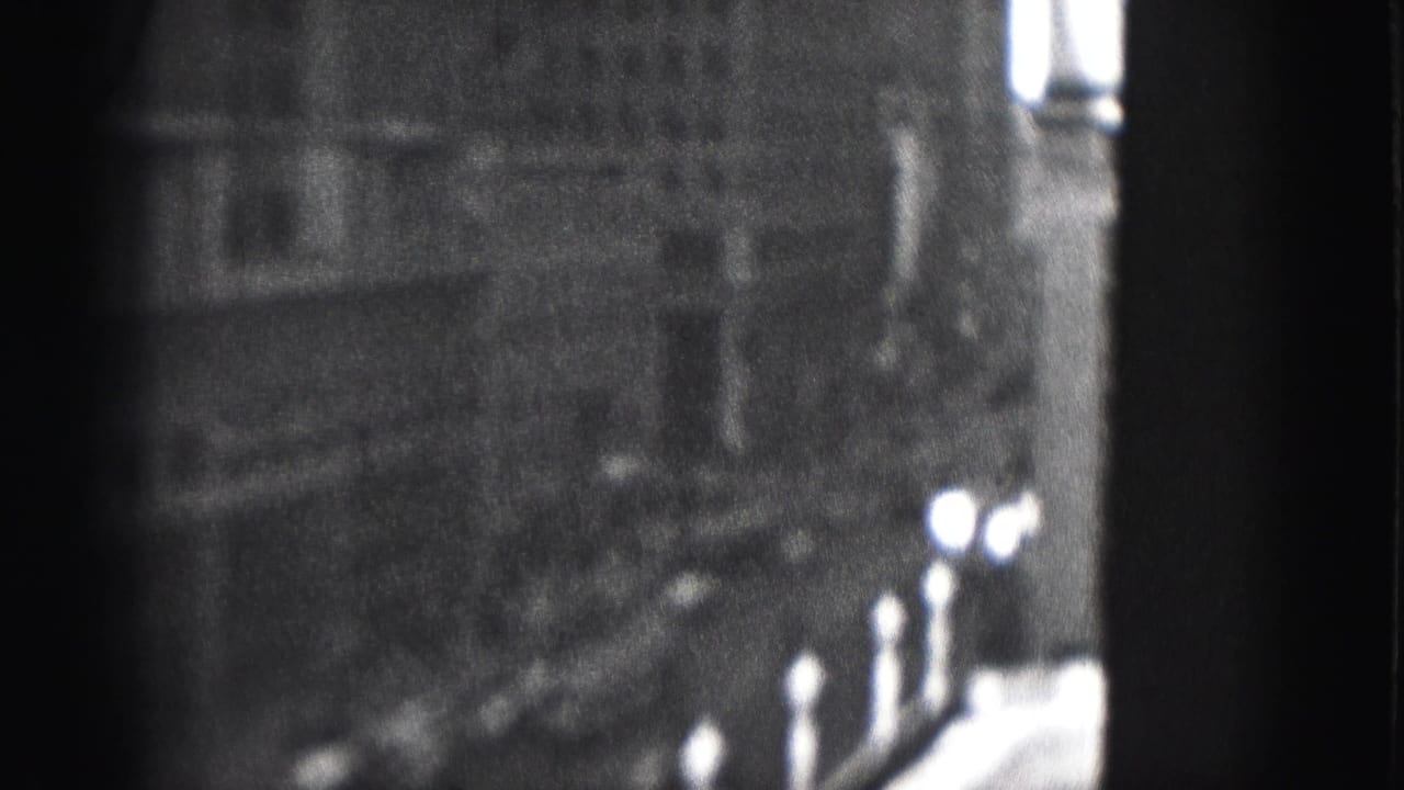 Grainy 1946 Harrisburg 8mm frame showing tall city buildings, sidewalk lampposts, and a shadowed street edge.