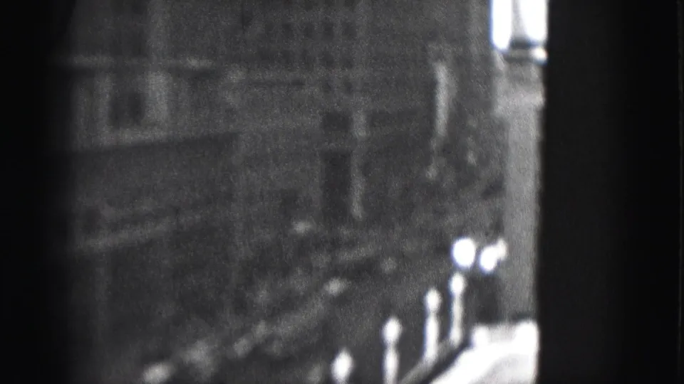 Grainy 1946 Harrisburg 8mm frame showing tall city buildings, sidewalk lampposts, and a shadowed street edge.