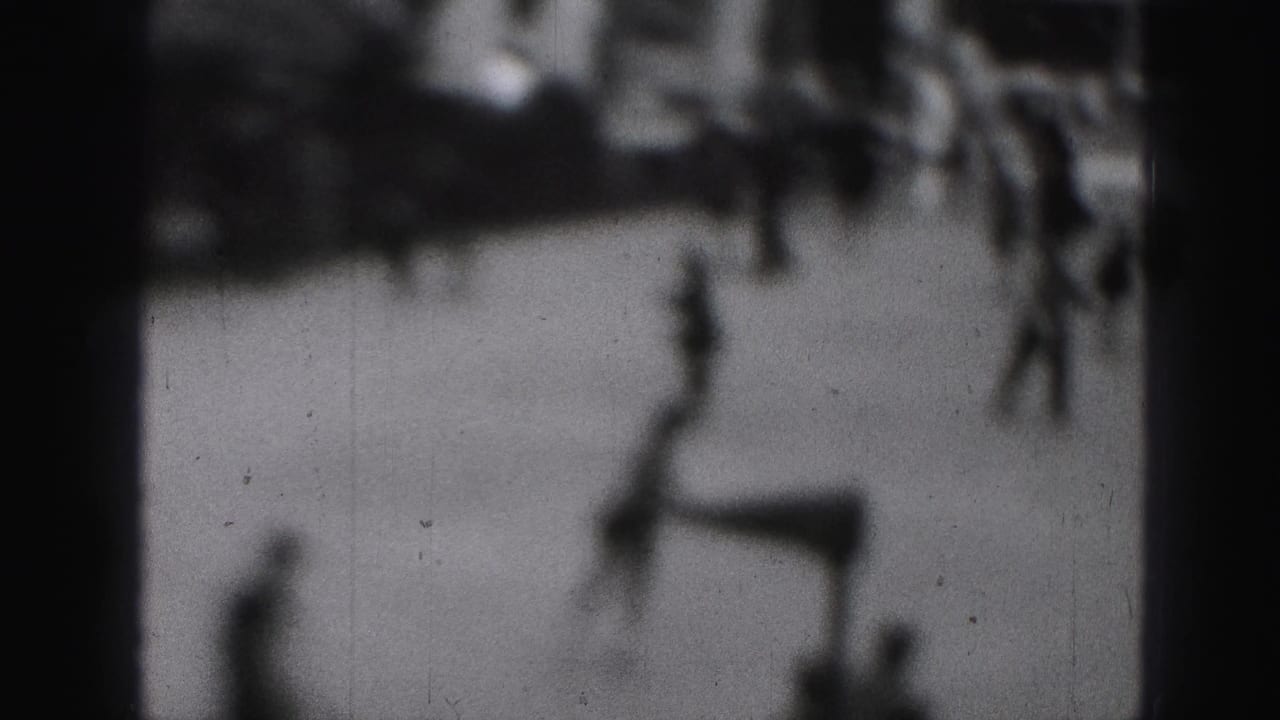 Blurred branches or figures pass in front of a pale background with dark film edges.