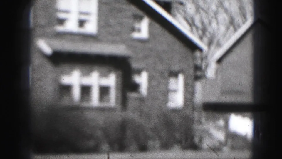 Black-and-white 8mm frame of a Harrisburg brick residence with windows, shrubs, and a partial roofline nearby.