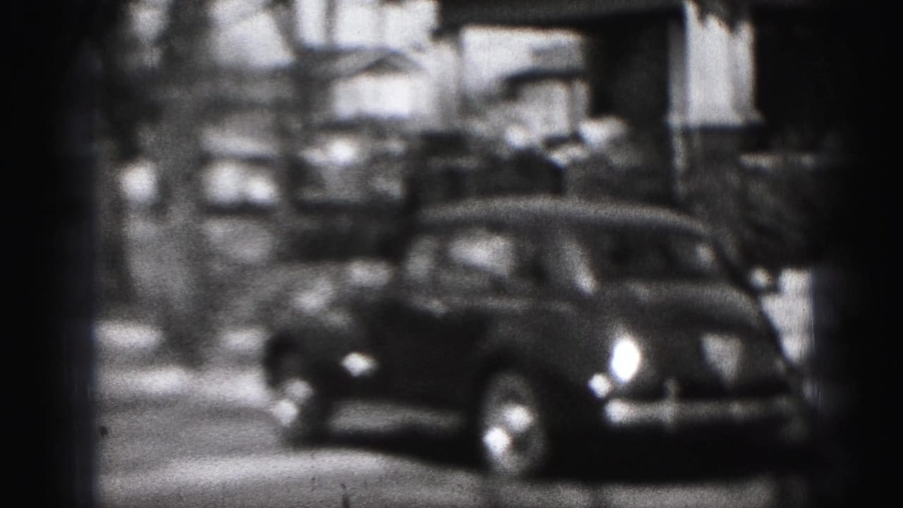 Parked rounded car on a residential street with houses and trees behind it.