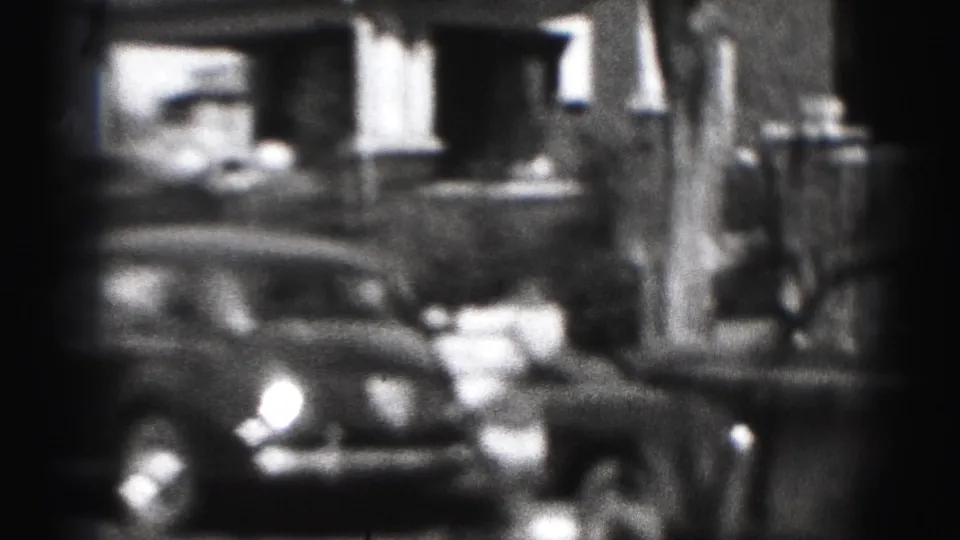 Black-and-white 8mm frame from Harrisburg showing a parked classic car near houses and a large tree trunk.