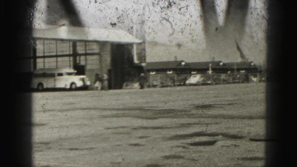 Home-movie frame showing a paved airfield or industrial area near Harrisburg, with vehicles, a hangar-like building, and distant people.