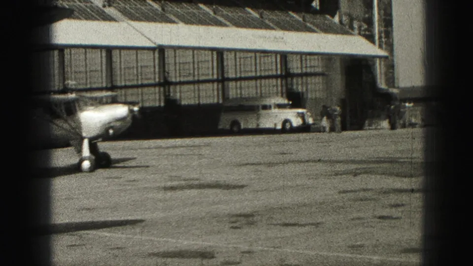 1946 Harrisburg frame showing a small aircraft on a paved area beside a hangar-like building, with vehicles and distant figures nearby.