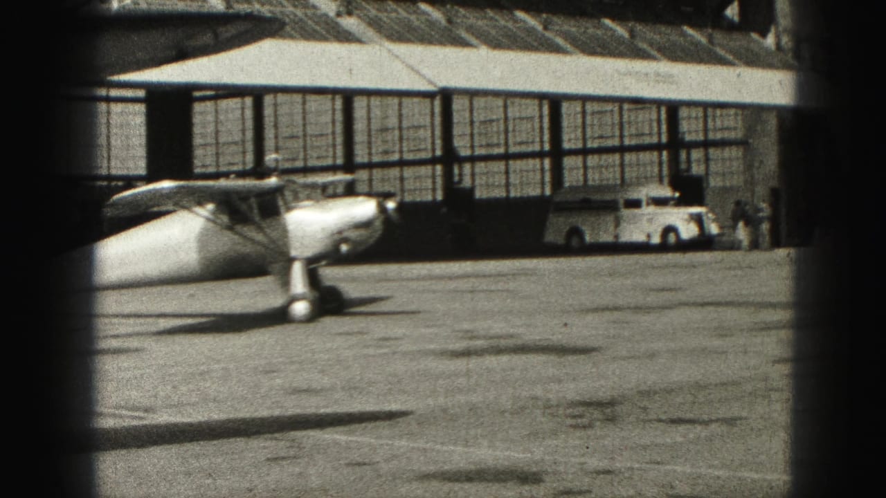 1946 Harrisburg home-movie frame showing a small aircraft near an airport hangar and a parked white car on the tarmac.