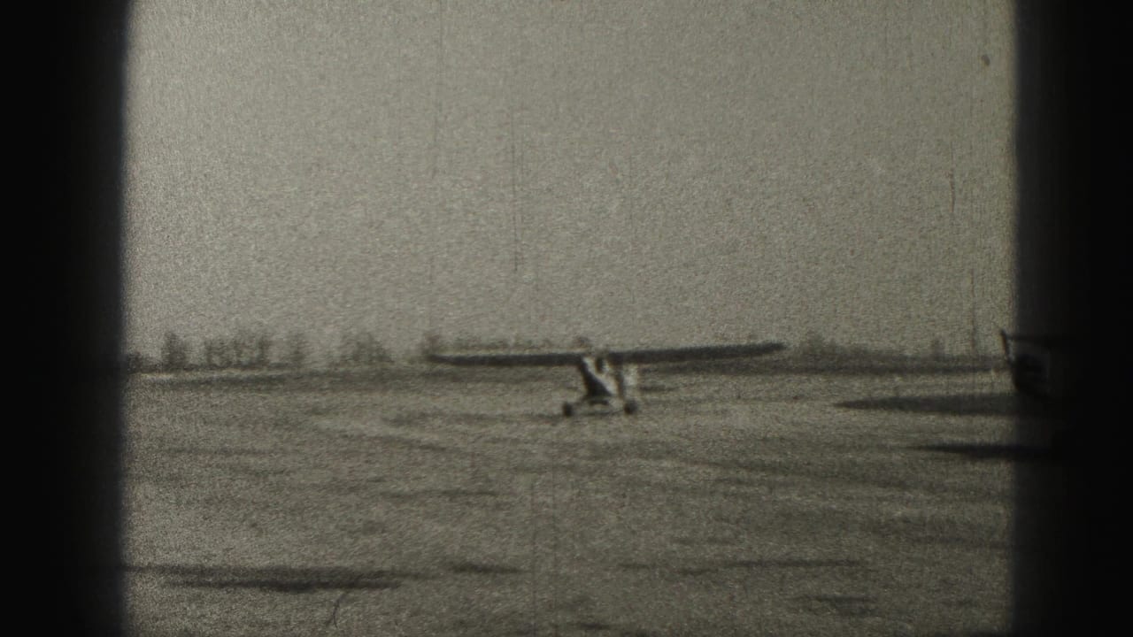 Small high-wing propeller plane taxiing across a flat airfield, seen through dark 8mm frame borders.