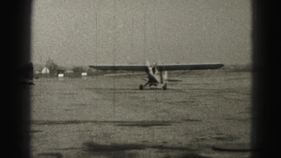 The plane nears the edge of the 1946 Harrisburg airfield view, with low distant structures and visible vertical film scratches in the frame.
