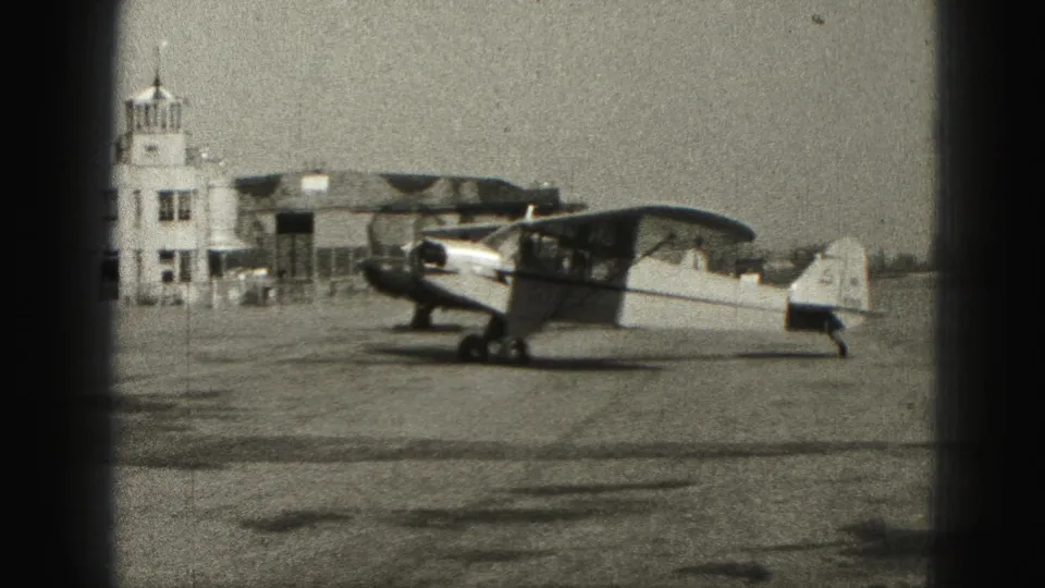Light aircraft parked on a Harrisburg airfield with an airport building and hangar-like structures in the background.