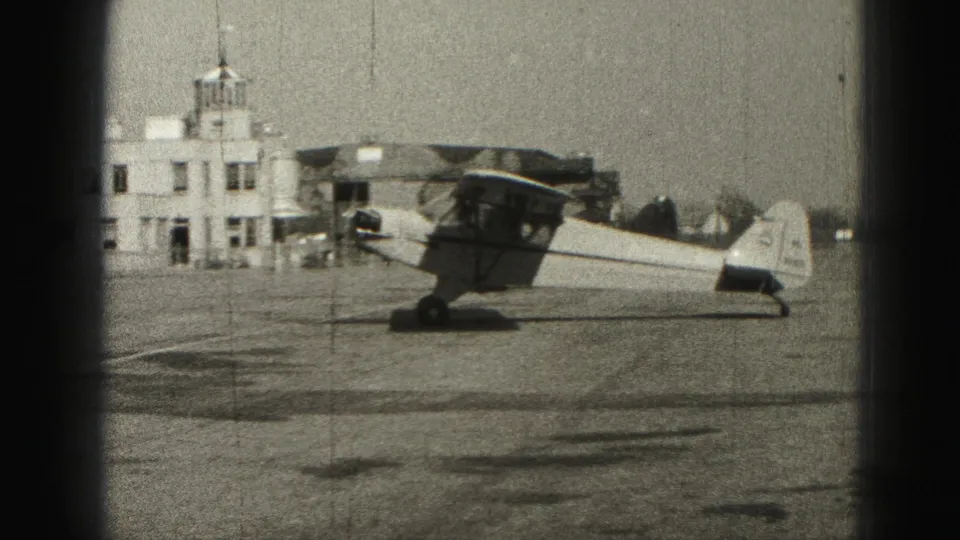 1946 Harrisburg-area 8mm frame showing a small high-wing aircraft on the tarmac near a white airfield building with a cupola.