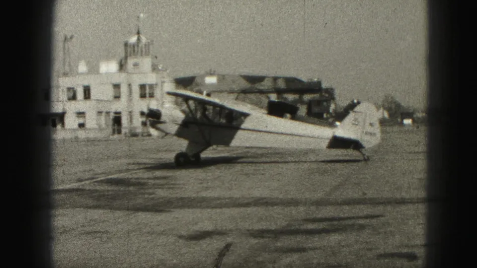Side view of a small single-engine aircraft at a Harrisburg airfield in 1946, with a low white building in the background.