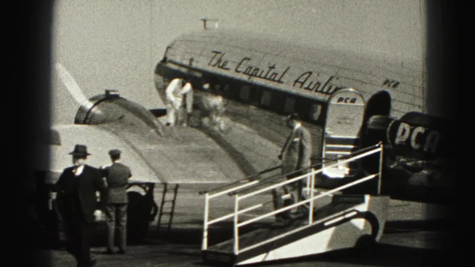 Two passengers in light clothing stand at the aircraft doorway as others wait around the stairway and airport apron.