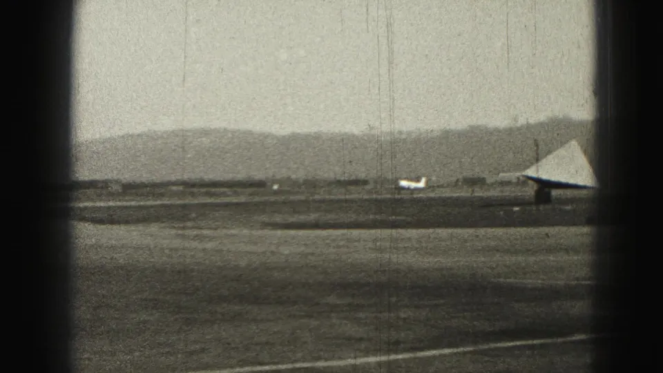 Small propeller aircraft continues along a Harrisburg runway in 1946, seen through soft black-and-white 8mm grain and gate borders.