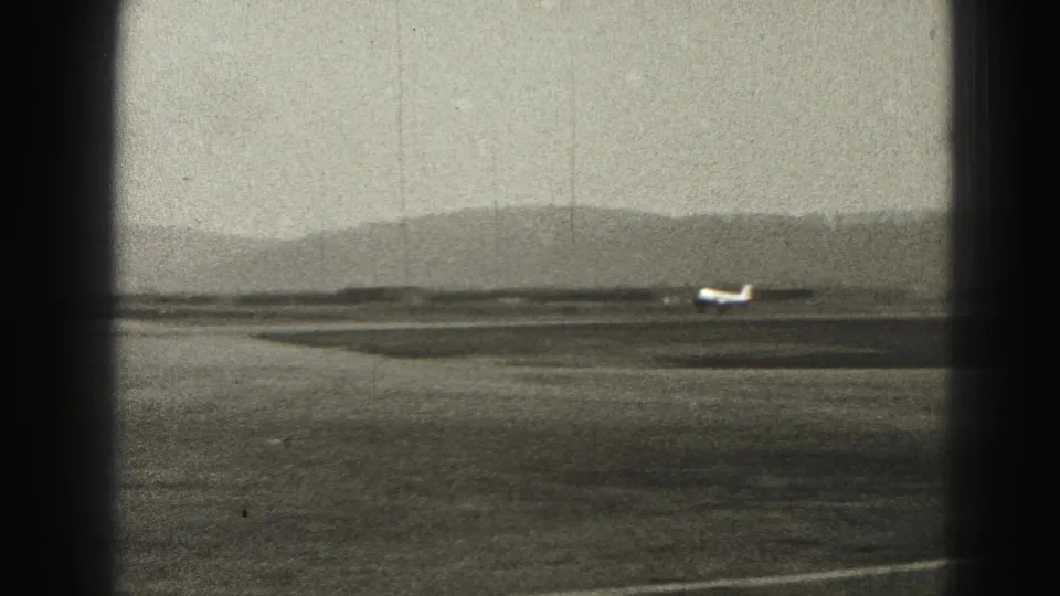 The small aircraft appears mid-run on the Harrisburg airfield, surrounded by open runway space and distant hills in a grainy 8mm frame.