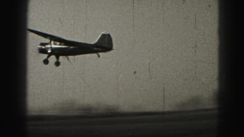 A small propeller plane crosses low in the frame, with soft 8mm grain and gate shadowing visible.
