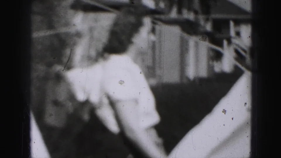Blurred adult woman in a light-colored dress appears near branches and vertical siding in 1946 Harrisburg home-movie footage.