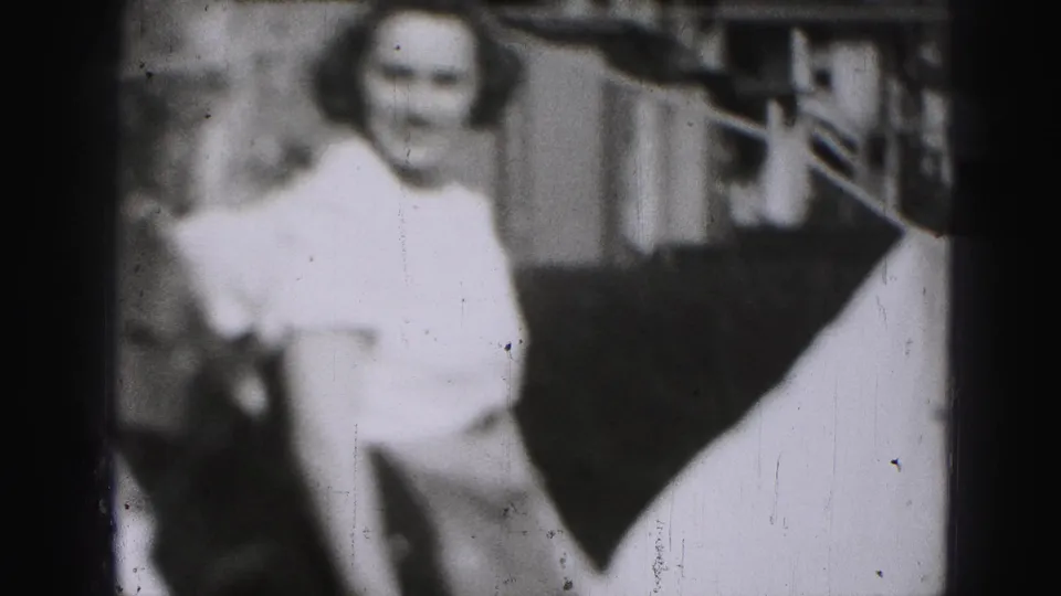 Adult woman, softly focused and wearing a light dress, stands near a building exterior in the Harrisburg 1946 home movie.