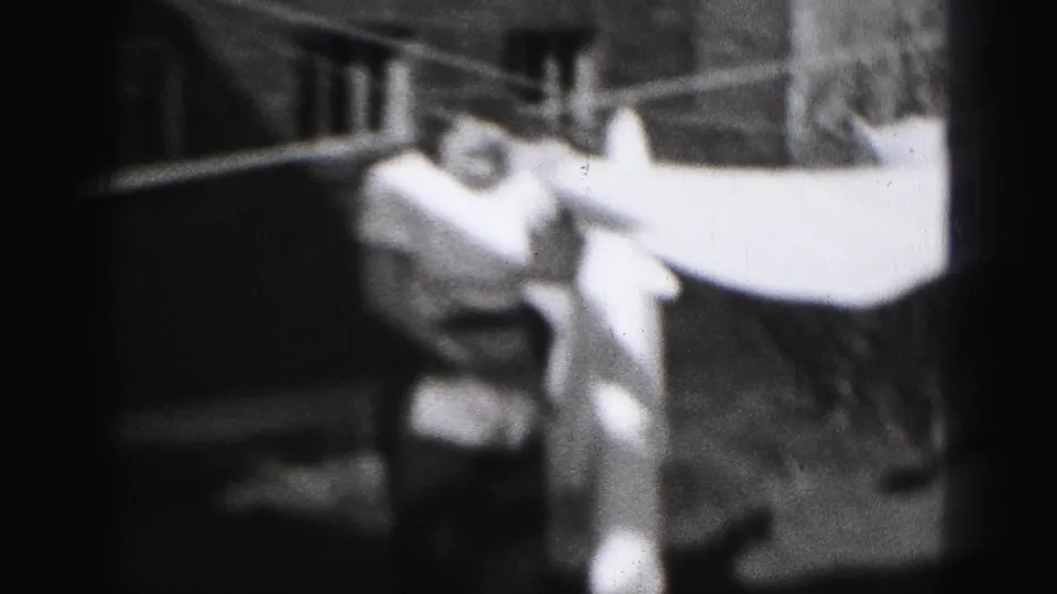 In Harrisburg, Pennsylvania, 1946, a woman in a light dress stretches a white sheet across an outdoor clothesline.
