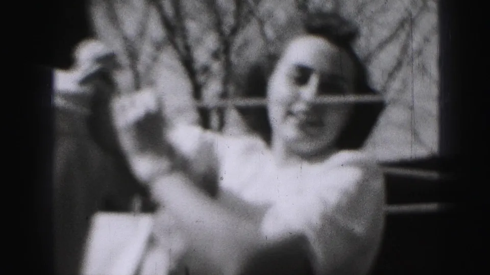 In 1946 Harrisburg home-movie footage, a woman in a light dress lifts laundry beside a backyard clothesline with bare branches behind her.
