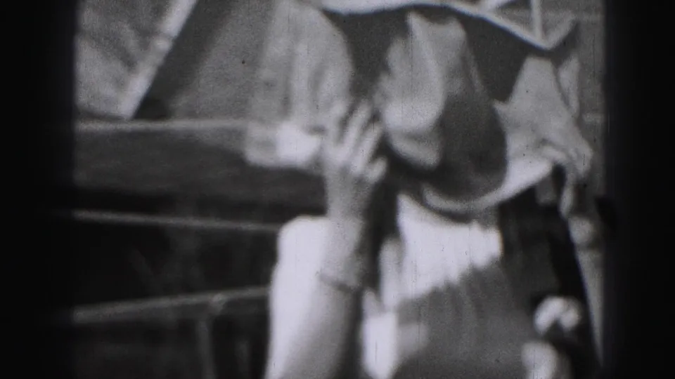A woman in period clothing stands close to the camera near hanging laundry, with outdoor steps or yard features behind her.