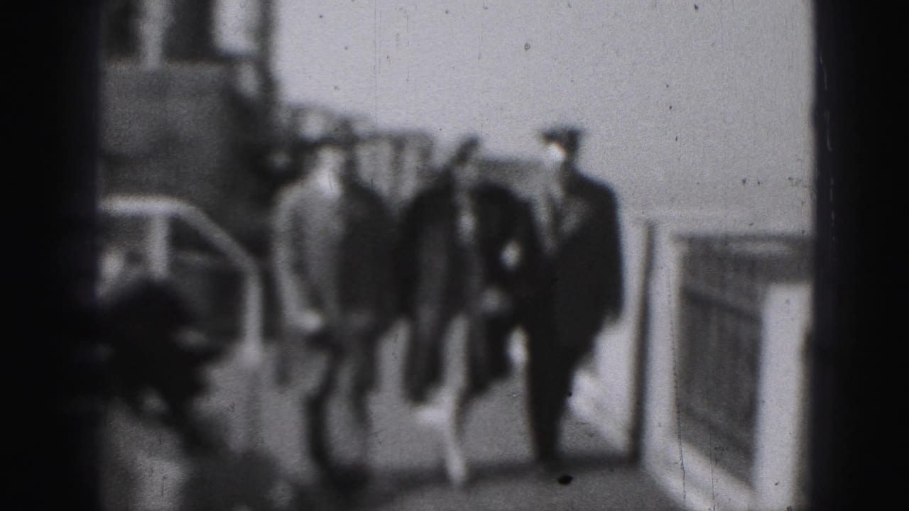 Three adults walk along a paved walkway beside railings, with a uniformed officer visible at right.