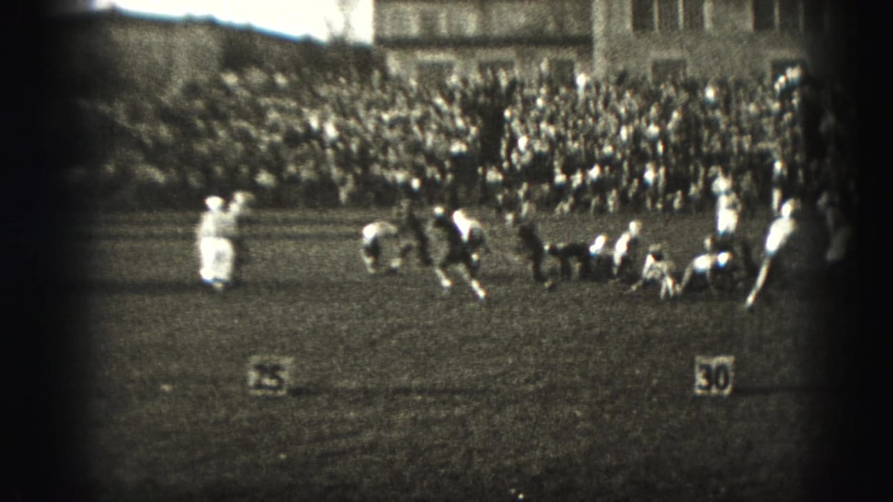 Football players crouch and move at the line of scrimmage with spectators behind the field.