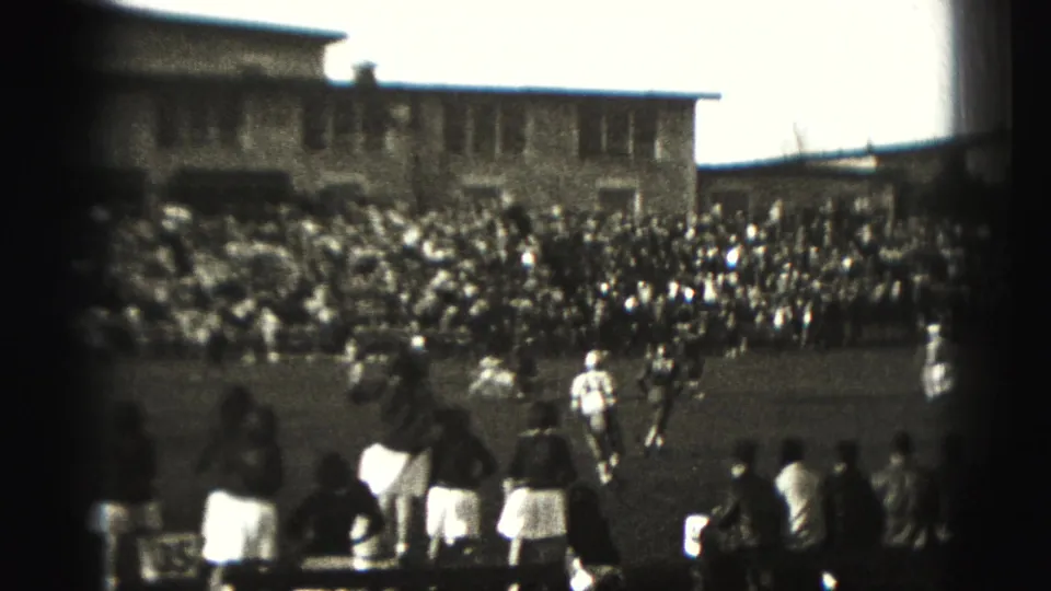 Sideline spectators and team figures watch a 1946 Harrisburg football game, with players and crowded bleachers in the background.