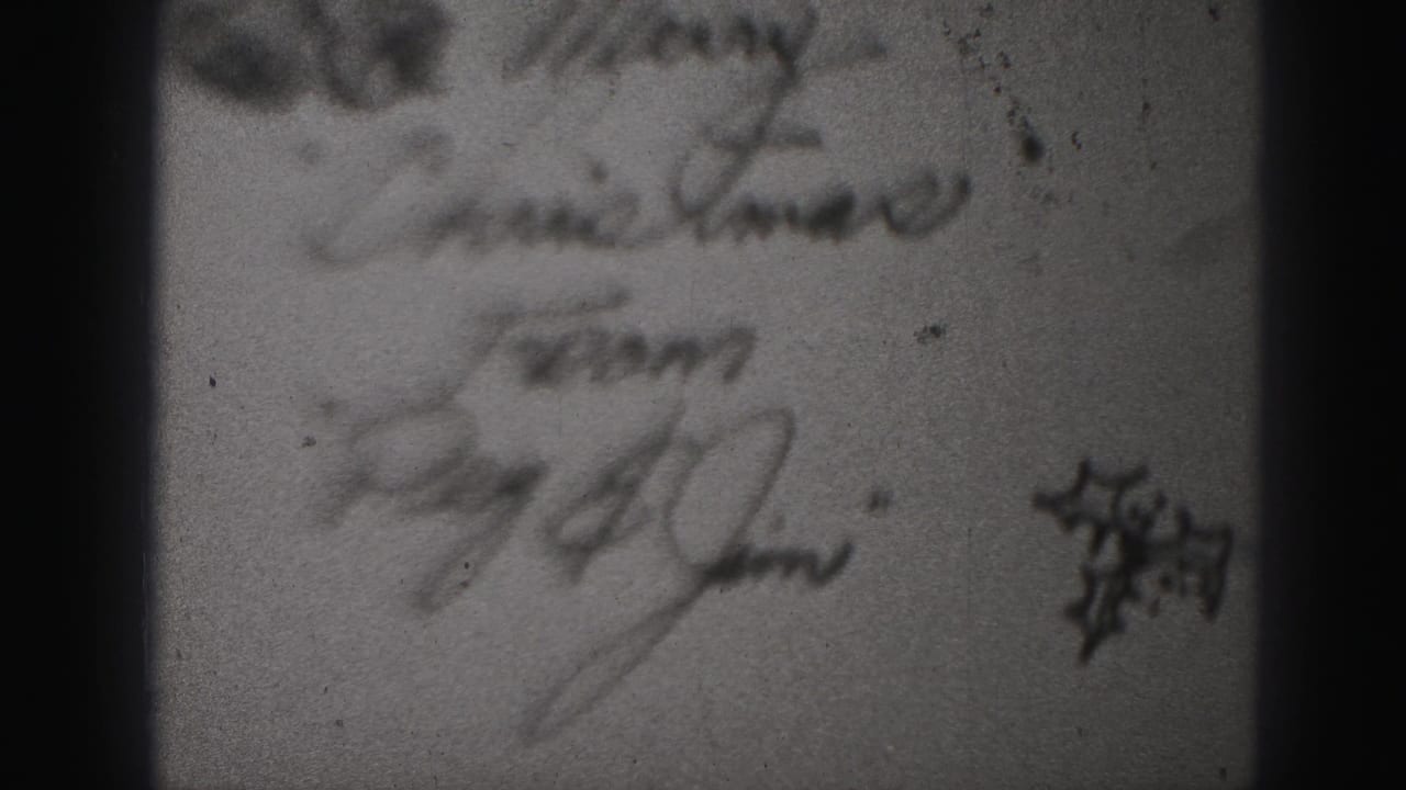 Close 8mm view of cursive writing on a paper item, with a small dark graphic at right and heavy softness from the film capture.
