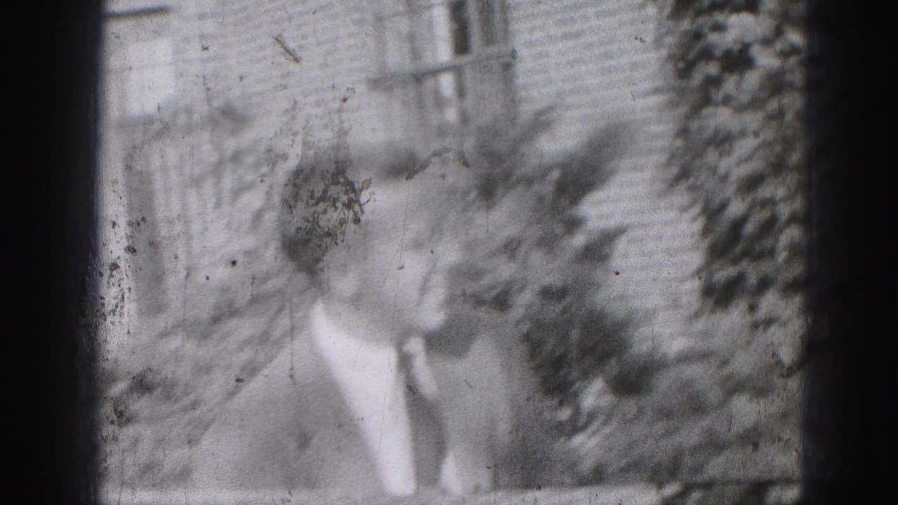 1946 Harrisburg home-movie frame showing a suited adult man outdoors, partly blurred, with brickwork, windows, and foliage behind him.