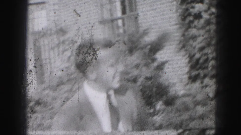 1946 Harrisburg home-movie frame showing a suited adult man outdoors, partly blurred, with brickwork, windows, and foliage behind him.