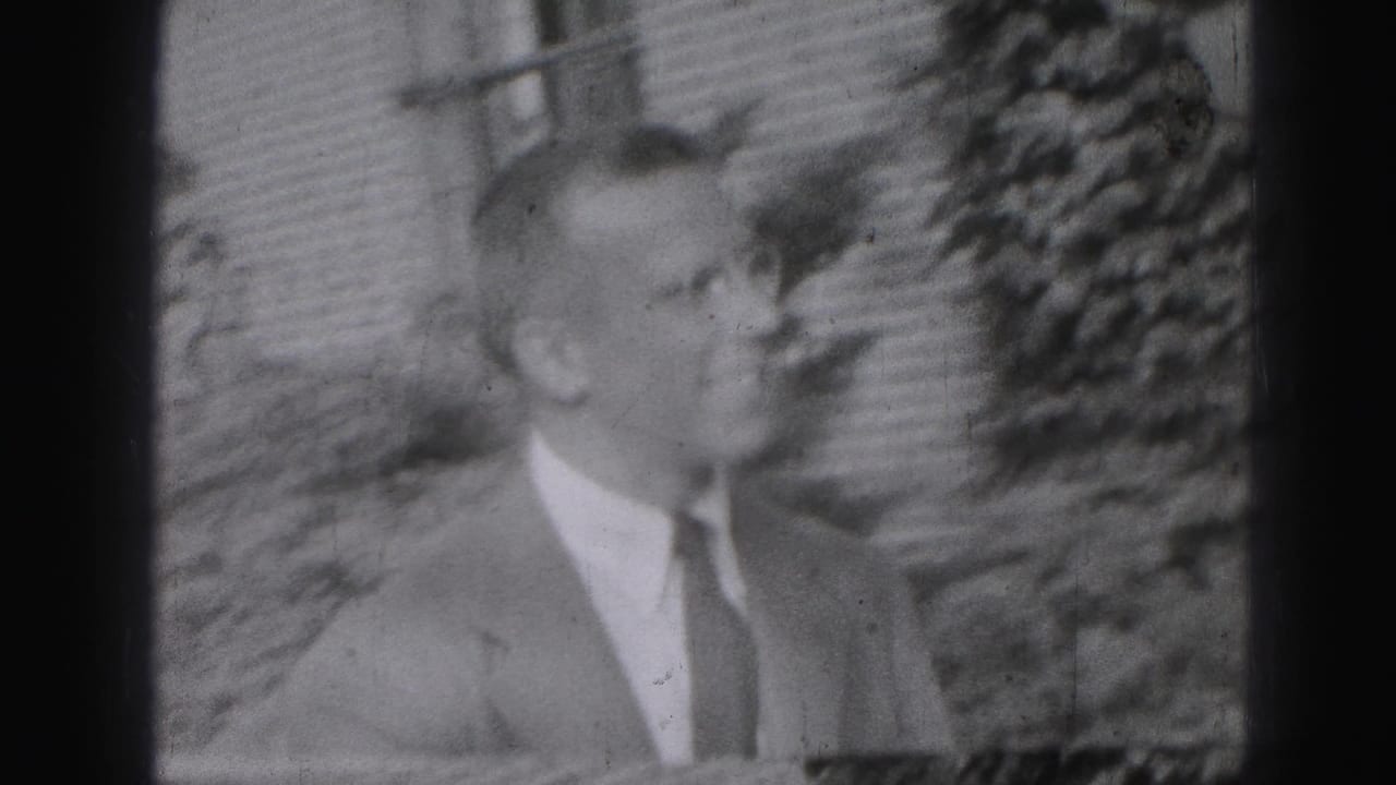 Suited man in profile outdoors, facing another person at the right edge of the frame.