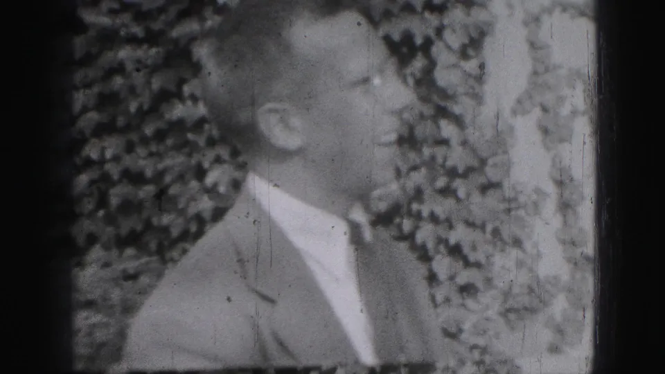 1946 Harrisburg home-movie frame showing a suited adult man smiling or speaking outdoors with dense foliage behind him.