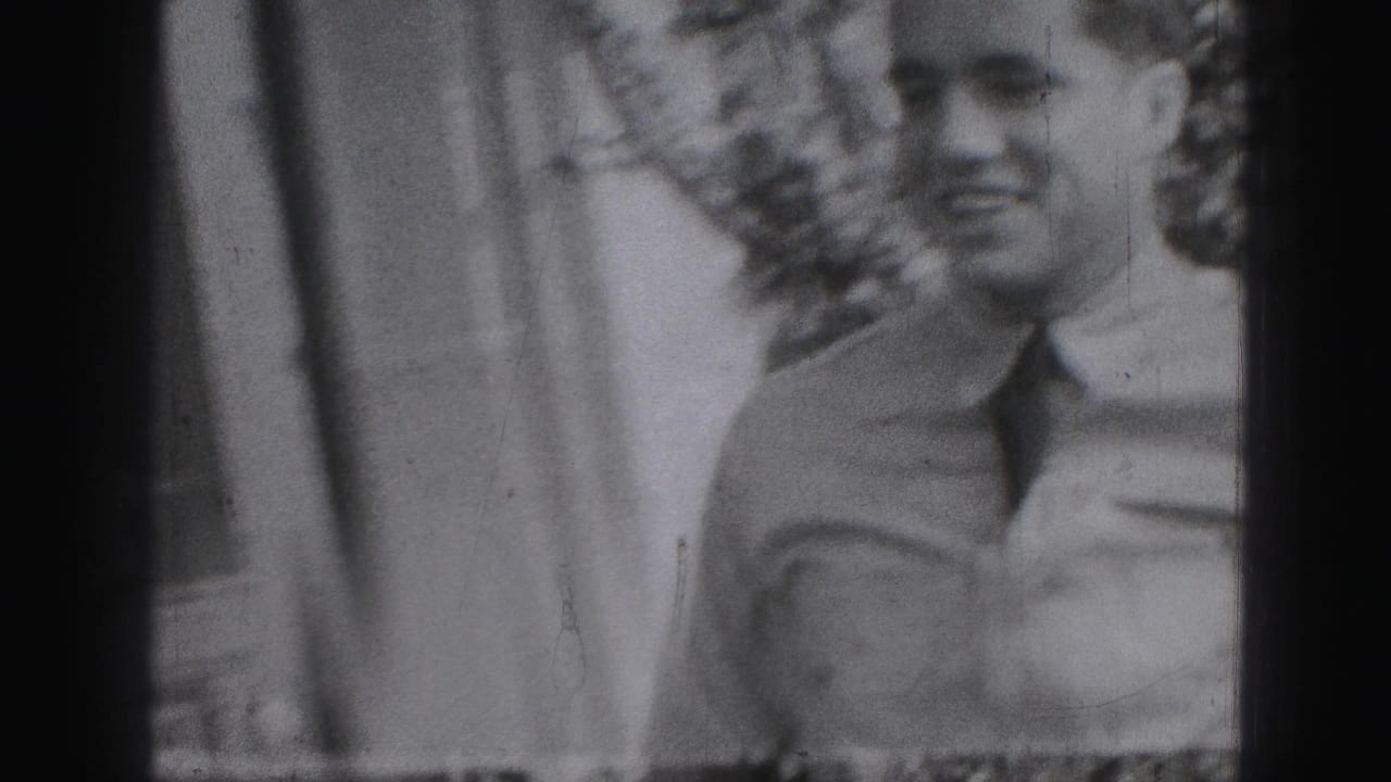 Man in a light shirt and tie stands near a house exterior in soft monochrome film.
