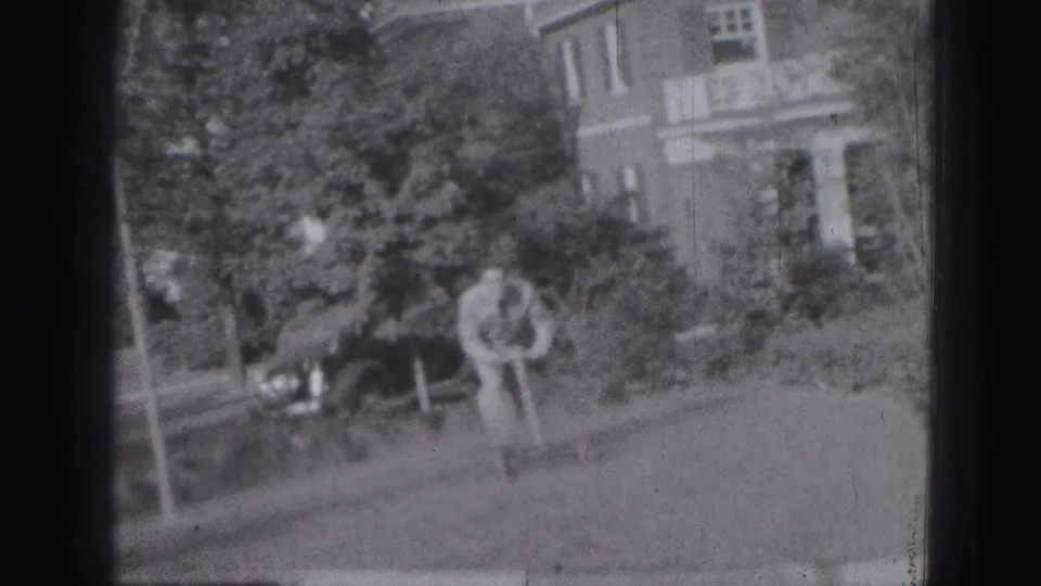 A man in 1946 Harrisburg yard footage bends near a small toy vehicle, with shrubs, a porch, and a parked dark car in the background.